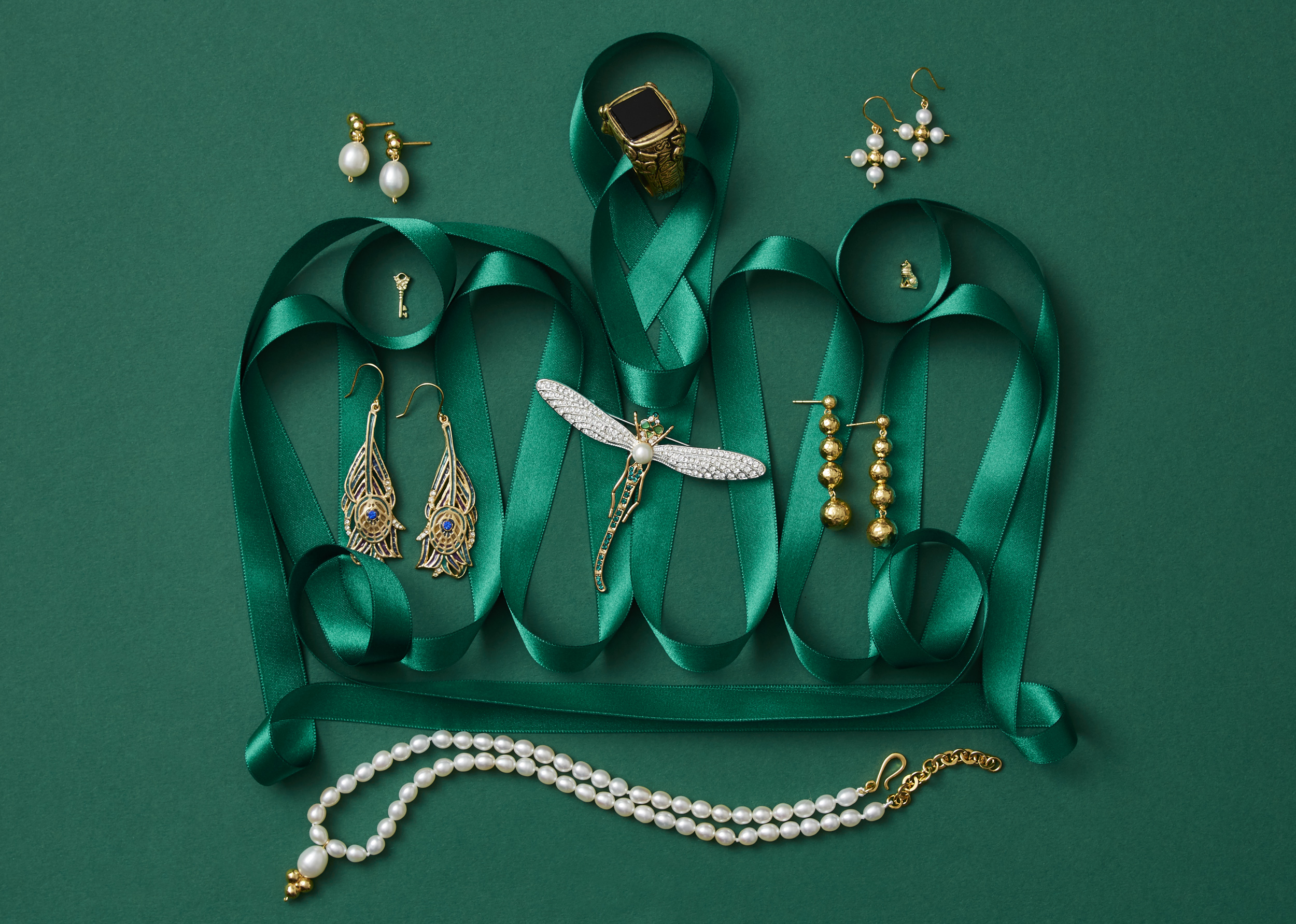 marketing still life photograph of a crown made of green ribbon adorned with gold and pearl jewelry including earrings, a pin, a ring, and a necklace on a green background.