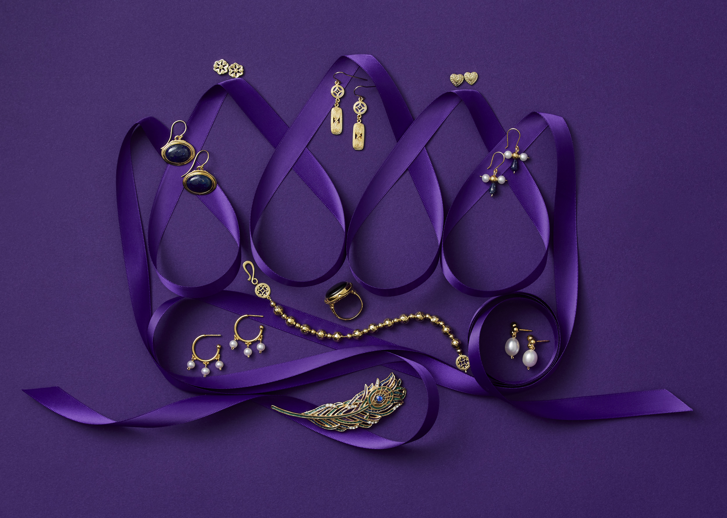 marketing still life photograph of a crown made of purple ribbon adorned with gold and pearl jewelry including earrings, a pin, a ring, and bracelets on a purple background.