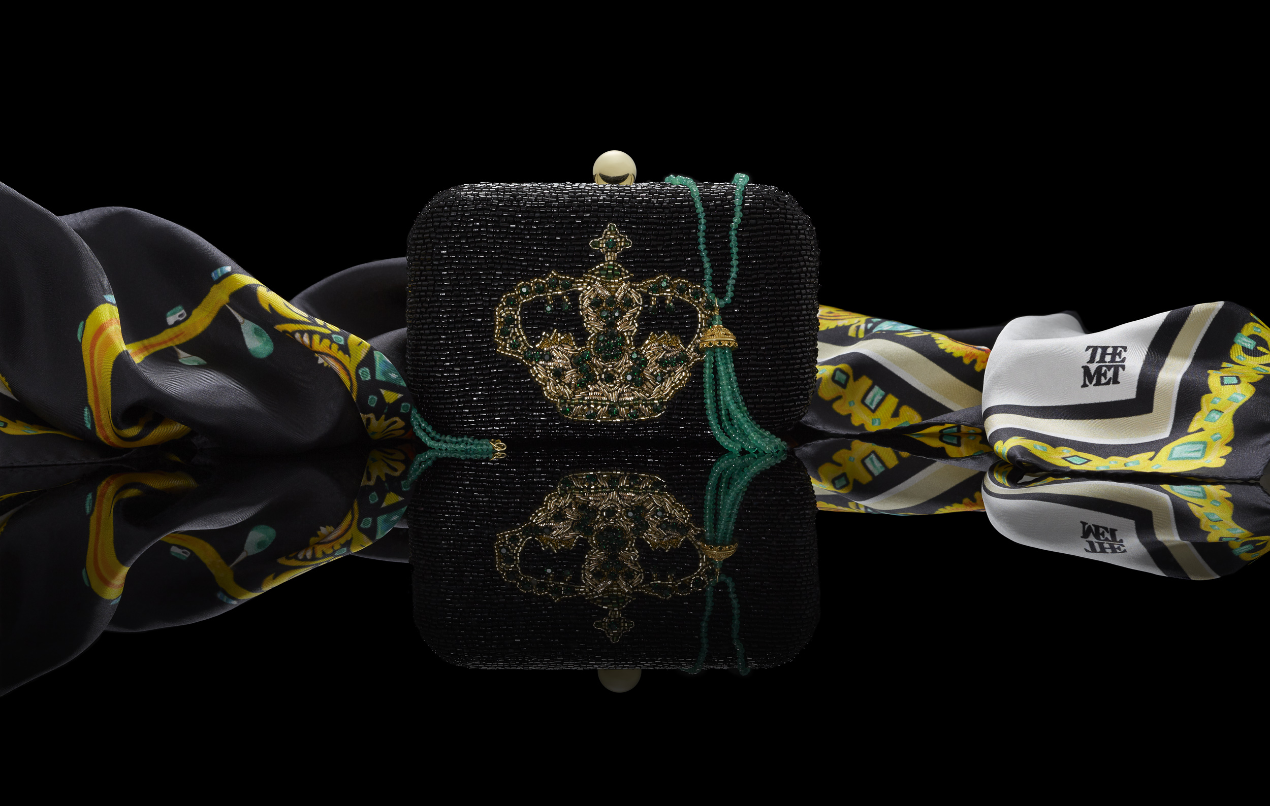 marketing still life photograph of the crown of the andes silk scarf, beaded clutch, and emerald necklace with reflection on a black background