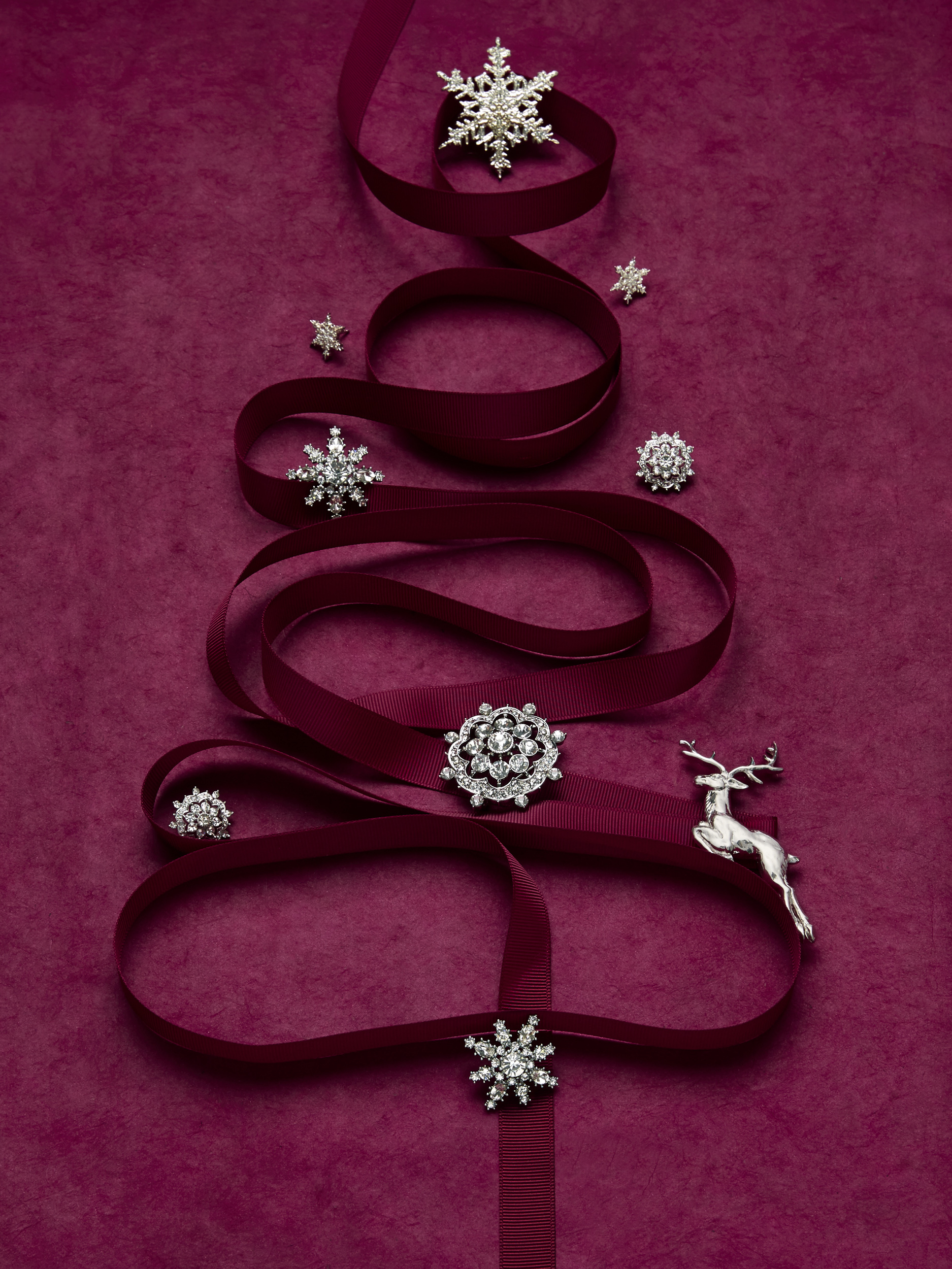 marketing still life photograph of silver and diamond jewelry including pins and earrings decorating a tree made of burgundy ribbon on a burgundy background