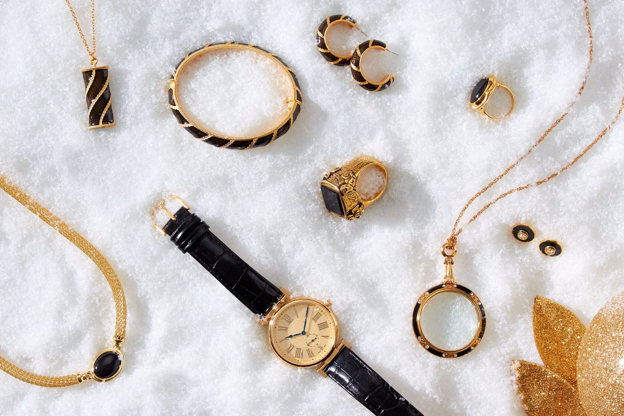 marketing still life photograph of black, gold and onyx jewelry including necklaces, earrings, rings, a bracelet, and a watch on snow