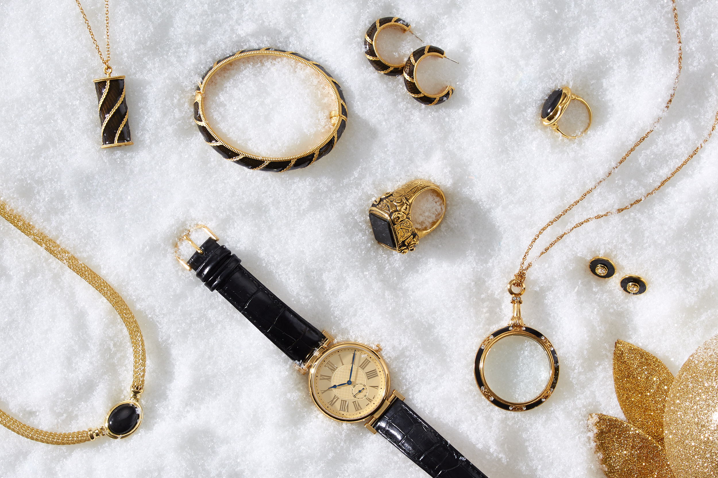 marketing still life photograph of black, gold and onyx jewelry including necklaces, earrings, rings, a bracelet, and a watch on snow