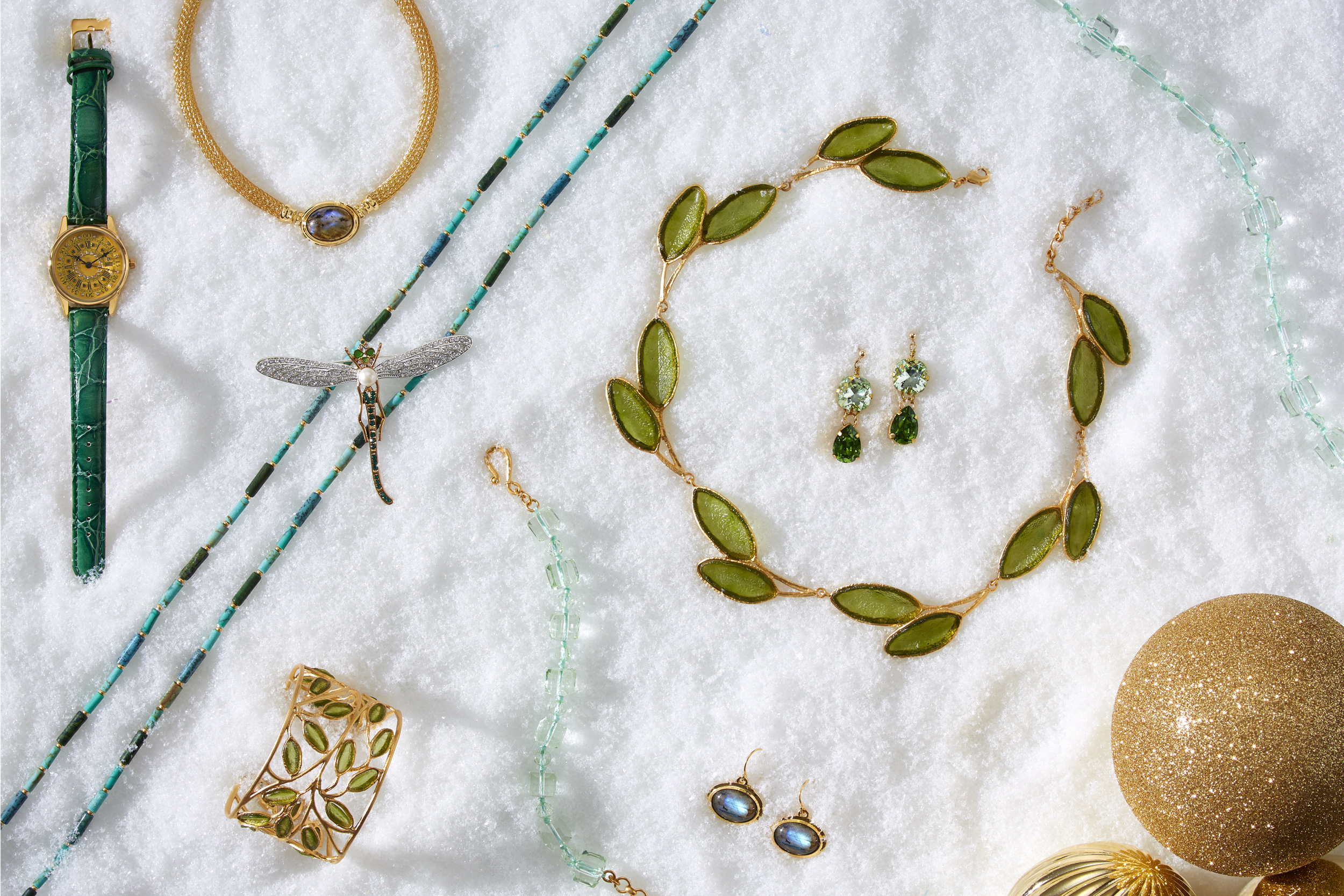 marketing still life photograph of green, gold, and turquoise jewelry including necklaces, earrings, bracelets, a watch, and a pin on snow