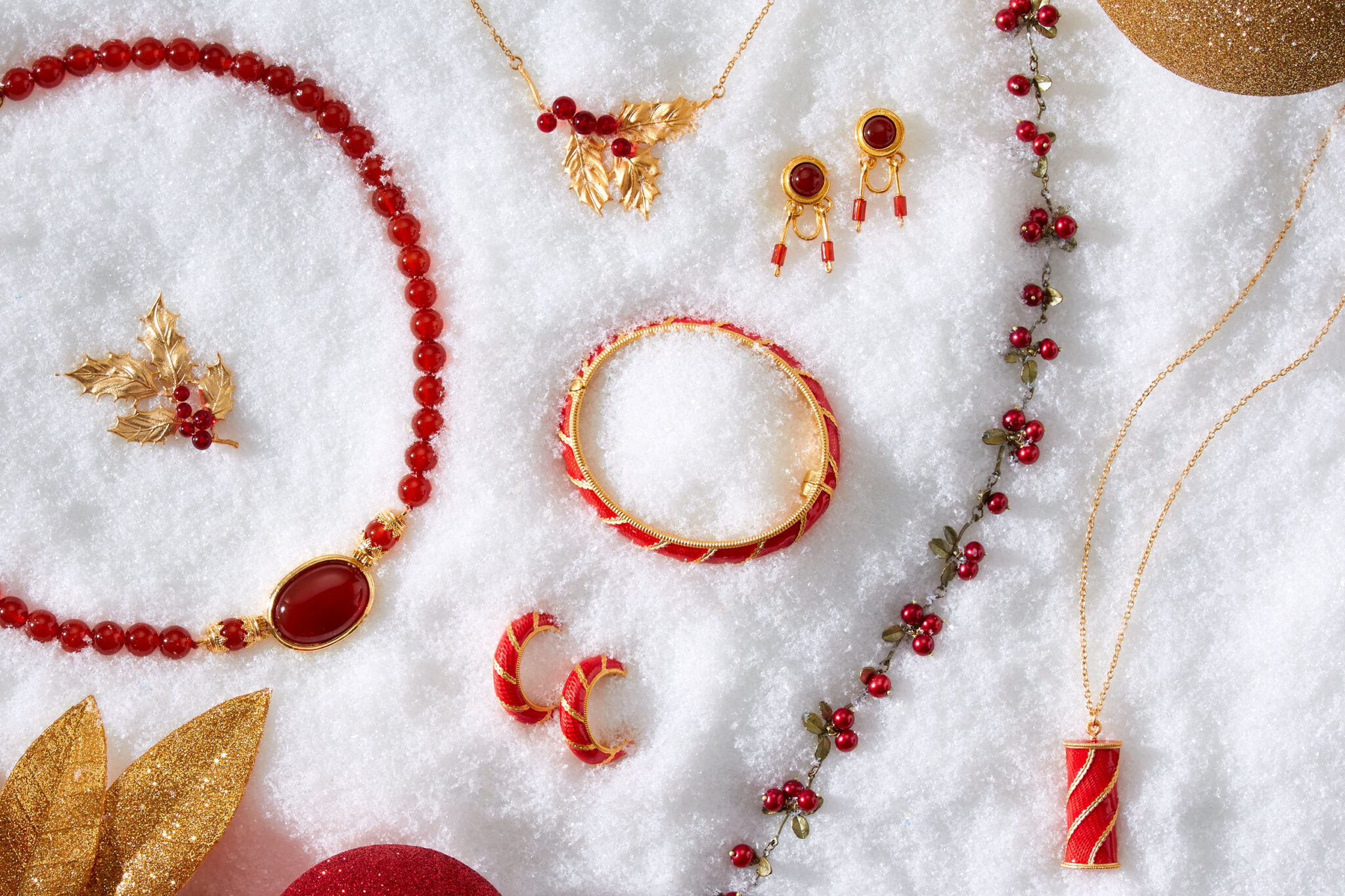 marketing still life photograph of red, gold, and garnet jewelry including necklaces, earrings, a bracelet, and a pin on snow