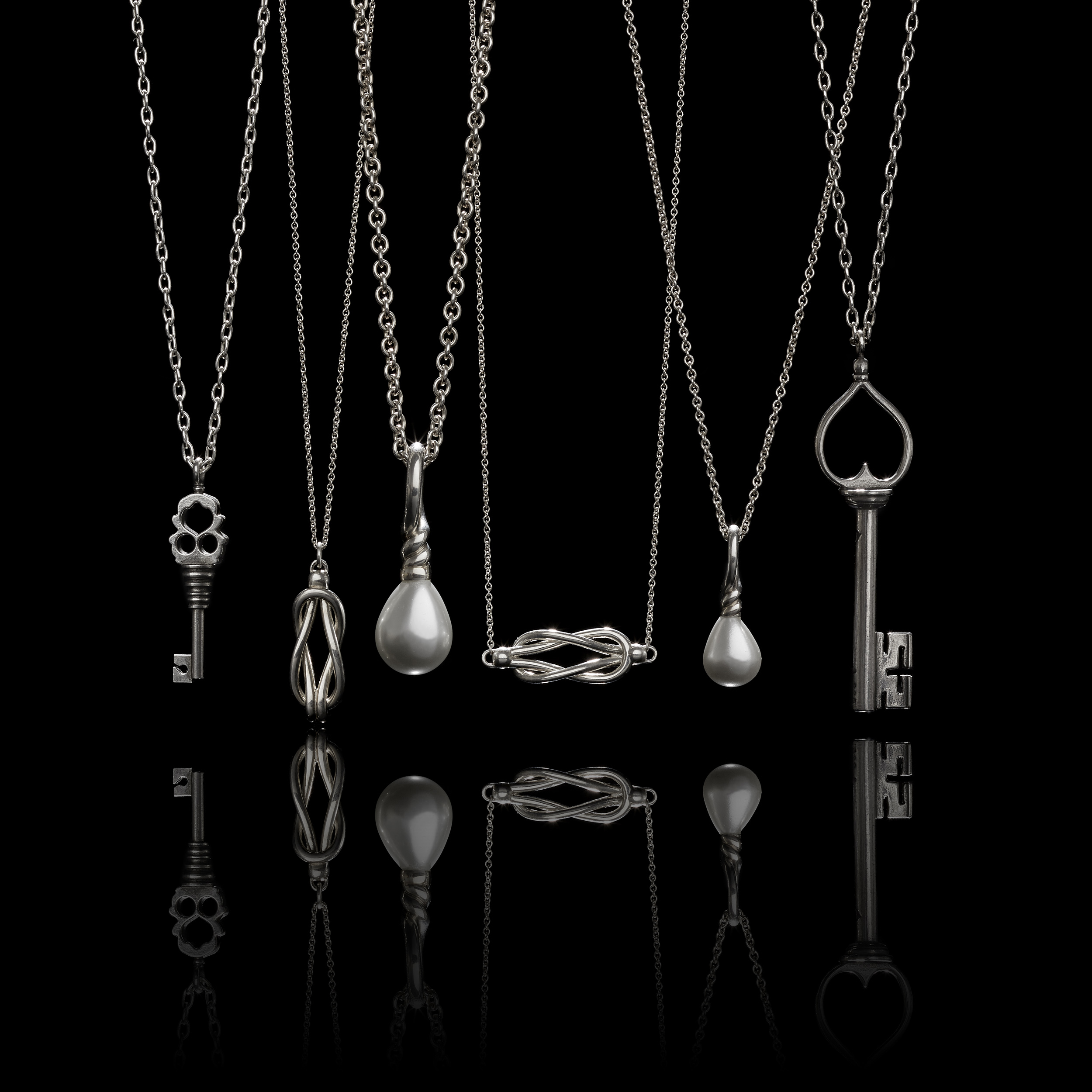 A group of silver pendant necklaces including keys, pearls, and knots with moody lighting, a reflection, and a black background