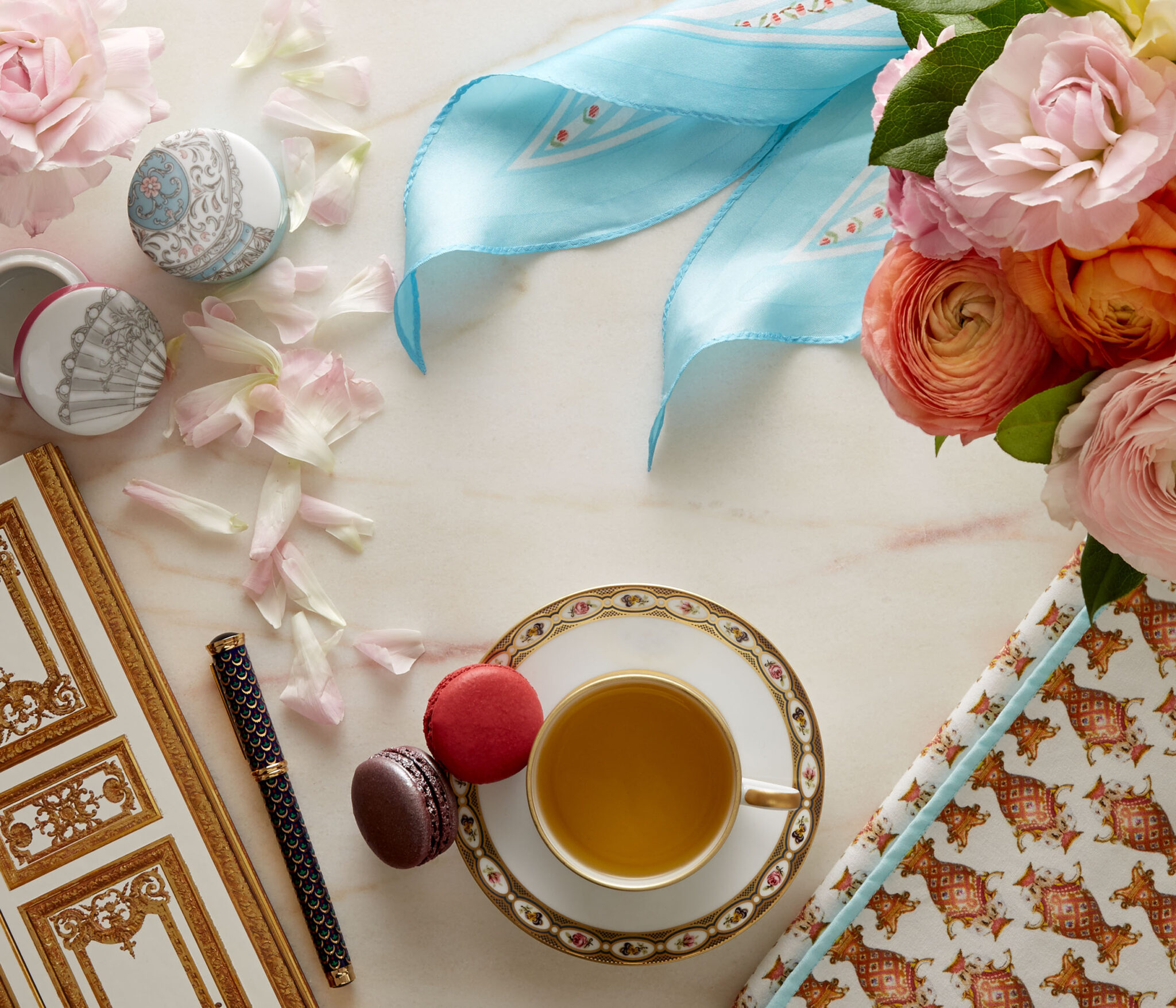still life in warm window light of a journal and pen, a scarf, macrons, tea, a napkin, and flowers on marble